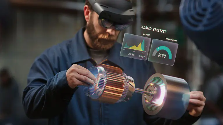 A technician wearing a HoloLens headset inspects a three dimensional model of an electric motor, with a systems check showing performance data floating beside it
