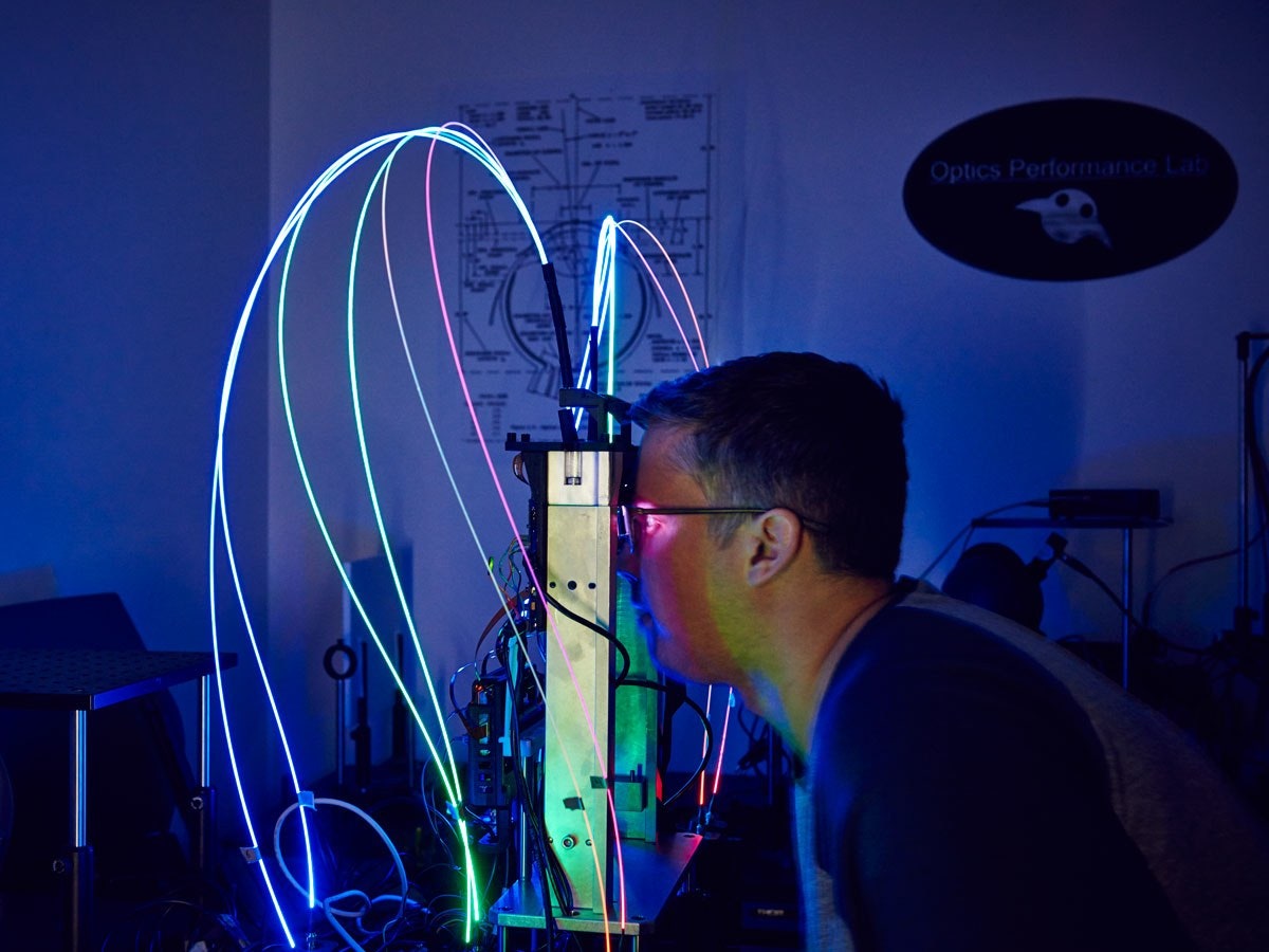 Eric Browy leaning toward a Magic Leap prototype in the Optics Performance Lab, with glowing fiber optic wires arching above it in the dark